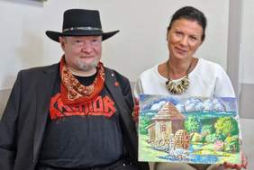 Pokaż zdjęcie: Pani Dyrektor w towarzystwie artysty Stanisława Kmiecika, znanego z niezwykłej techniki malowania przy użyciu nóg. Pani Dyrektor trzyma w dłoniach barwny obraz autorstwa artysty, przedstawiający drewniany młyn wodny w otoczeniu bujnej zieleni i błękitnego nieba z obłokami. Obie postacie uśmiechają się, prezentując dzieło sztuki.