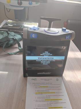 Pokaż zdjęcie: Zdjęcie przedstawia urządzenie omniReader, które służy do odczytywania tekstu na głos dla osób niewidomych i słabowidzących. Na ekranie urządzenia widoczne jest menu ustawień. Na stole obok leży kartka z logo OWiT (Ośrodek Wsparcia i Testów) w Białymstoku, zawierająca informacje o realizacji zadań w ramach działalności ośrodka. W tle znajduje się inne urządzenie wspomagające.