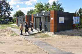 Pokaż zdjęcie: Zdjęcie kontenerów mieszkalnych przed nimi stoją ludzie, część osób zagląda do środka
