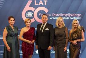Pokaż zdjęcie: Cztery elegancko ubrane kobiety i jeden mężczyzna w garniturze wspólnie pozują do zdjęcia podczas gali. W tle granatowa ścianka z napisem „60 LAT Sportu Paralimpijskiego w Województwie Podlaskim” i logo „START”. Zdjęcie oficjalne, pamiątkowe.
