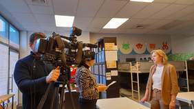 Pokaż zdjęcie: Po lewej stronie zdjęcia stoi operator kamery, ujęty w momencie filmowania. Kamera skierowana jest na dwie rozmawiające kobiety. Jedna z kobiet, uczesana jest kucyk, ubrana w czarno białą marynarkę i czarne spodnie, druga z pań blondynka, ubrana jest w beżowy garnitur i białą bluzkę 