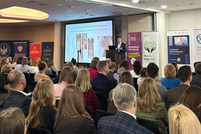 Pokaż zdjęcie: Uczestnicy konferencji siedzą w sali podczas prelekcji. Na scenie prelegent przy mównicy, za nim ekran z prezentacją o skoliozie oraz banery partnerów wydarzenia „Oswoić skoliozę”.