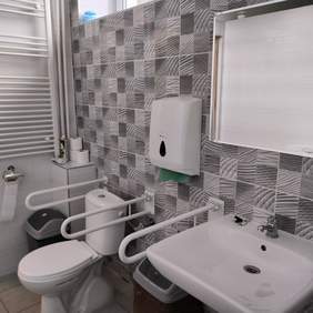 Pokaż zdjęcie: Zdjęcie jednej ze ścian w łazience. Na ścianie przyklejona jest szaro biała wzorzysta glazura. Na zdjęciu widać sedes w kolorze białym z uchwytami, obok niego jest umywalka nad którą wisi lustro. Na ścianie zamontowany jest podajnik na papier. Po obydwu stronach sedesu stoją kosze plastikowe z przykrywkami. 