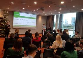 Pokaż zdjęcie: Grupa kilkunastu osób siedzi w sali konferencyjnej. Z przodu sali, po prawej stronie stoi przed mównicą kobieta, która trzyma w ręce mikrofon i przemawia do zgromadzonych osób. Na sali, po lewej stronie, przed udekorowaną bombkami choinką stoi kobieta, która jest tłumaczem języka migowego. W tle, na dużym ekranie wyświetlony jest plakat promujący konferencję pt. „Wizja - Wiedza - Wsparcie czyli WWW o dostępności przestrzeni społecznej”.