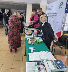 Pokaż zdjęcie: Na zdjęciu znajduje się stoisko informacyjne w budynku z kafelkową podłogą i sufitem z płytami kasetonowymi. Stoisko jest obsługiwane przez kilka osób, a na stolikach znajdują się różne materiały informacyjne, broszury, formularze, długopisy oraz inne gadżety promocyjne. Widoczny jest baner Państwowego Funduszu Rehabilitacji Osób Niepełnosprawnych (PFRON) z informacją o Centrach Informacyjno-Doradczych dla Osób z Niepełnosprawnością (CIDON). Przy stoisku znajdują się osoby, które rozmawiają z obsługą, a niektórzy z nich trzymają materiały w rękach. Atmosfera wydaje się profesjonalna, a całość przypomina targi lub konferencję tematyczną.