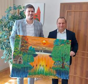 Pokaż zdjęcie: (Dwóch mężczyzn ubranych w białe koszule i marynarki trzyma w rękach kolorowy obraz. Znajdują się w pokoju, za nimi drzwi i kwiat