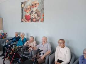 Pokaż zdjęcie: Na kanapie siedzą starsze panie. Niektóre trzymają przed sobą balkoniki. Kobiety uśmiechają się serdecznie. 