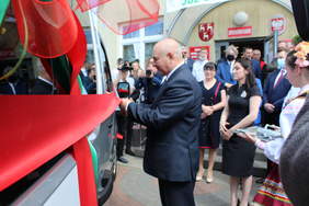 Pokaż zdjęcie: Mężczyzna trzyma w ręku nożyczki ,którymi przecina wstęgę ,która jest na nowym busie. W tle budynek  szkoły gdzie stoją mężczyżni i kobiety