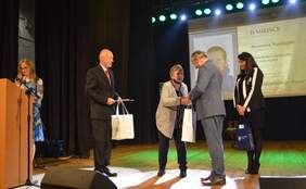 Pokaż zdjęcie: na scenie trzy osoby wręczają laureatce konkursu plastycznego nagrody w tle widać zdjęcia nagrodzonej pracy