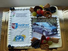 Pokaż zdjęcie: Kwadratowy tort w kolorze białym kolorze, na środku tortu grafika z autem.