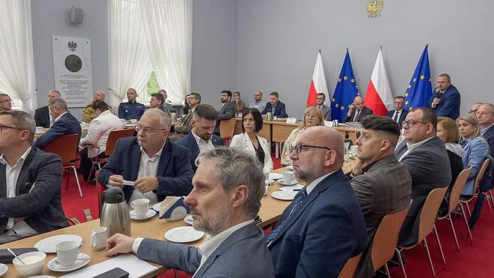 Na sali, przy stołąch siedzą uczestnicy spotkanai - kobiety i mężczyźni. W tle stoją flagi Polski i Unii Europejskiej