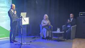 Pokaż zdjęcie: Konferansjer stoi przy mównicy i mówi do siedzących obok na fotelach pracowników PFRON