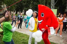 Pokaż zdjęcie: Do maskotki symbolizującej kroplę krwi przytula się do zdjęcia dziewczyna pokazując kciuk w górę. Maskotka trzyma flagi polskie i balony.