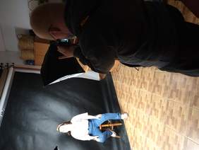 Pokaż zdjęcie: Mężczyzna w okularach trzymający profesjonalny aparat fotograficzny przymierza się do zdjęcia. Na stołeczku pozuje do zdjęcia dziewczynka z zespołem downa. Od modelki odbijają się światła reflektorów.