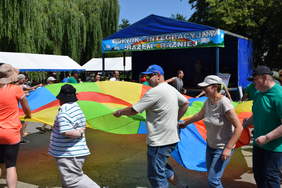 Pokaż zdjęcie: Grupa kilku osób trzyma kolorowy materiał w formie koła, zabawa na świeżym powietrzu