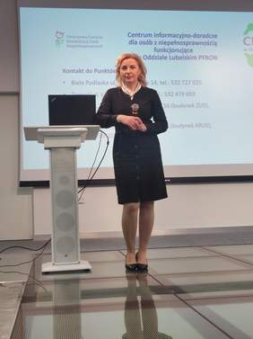 Pokaż zdjęcie: Kobieta blondynka w czarnej sukience stoi w lekkiej pozie oparta łokciem o mównicę, w ręku trzyma mikrofon