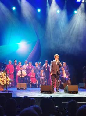 Pokaż zdjęcie: scena w Teatrze Muzycznym w Łodzi ,  na scenie światła w kolorze niebieskim, na scenie stoją artyści studia integracji i śpiewają, na pierwszym planie chłopak ubrany w piękny czarny garnitur trzyma mikrofon i śpiewa dla dużej publiczności 