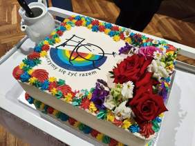 Pokaż zdjęcie: Kolorowy tort z tęczowymi warstwami. Na torcie znajduje się logo espołu Niepublicznych Szkół im. Alicji po drugiej stronie lustra oraz dekoracje z żywych kwiatów
