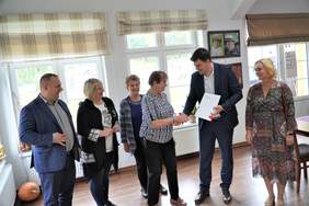 Pokaż zdjęcie: W świetlicy ośrodka PSONI. Pan starosta w kraciastym garniturze i koszuli w kratę. Obok pani w białej koszulce, jeansach i czarnej kurtce. Starsza kobieta i młody mężczyzna podający sobie ręce, za nimi pani w sweterku. Po prawej blondynka z sukie