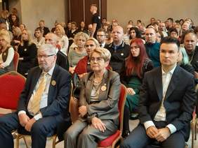 Pokaż zdjęcie: W sali znajdują się rzędy krzeseł. Siedzą na nich ludzie. Słuchają wykładów i prelekcji. 