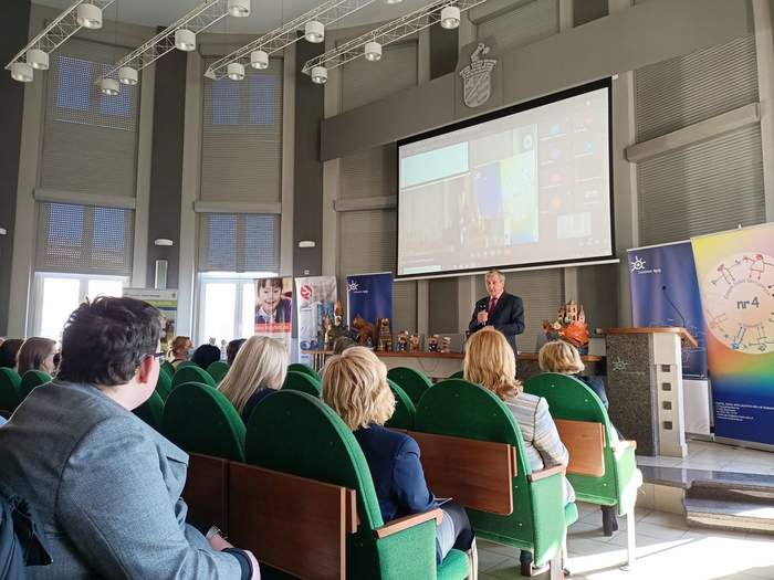 Pokaż zdjęcie: Fragment sali, na podwyższeniu stoi mężczyzna w garniturze i trzyma mikrofon. W tle na ścianie znajduje się duży ekran. W rzędach na fotelach siedzą ludzie.