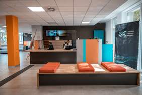 Pokaż zdjęcie: W sekretariacie za drewnianym blatem siedzi kobieta w okularach i jasnej bluzce. Pracuje na komputerze, trzyma myszkę w ręku myszkę. Po prawej stronie baner z ciemnym tłem na nim jasny napis Gdyńskie Centrum Kontaktu