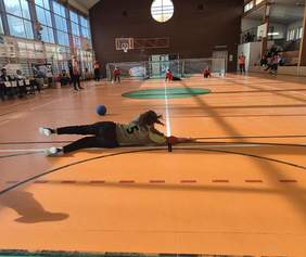 Zdjęcie przedstawia scenę z meczu goalball na hali sportowej z pomarańczową podłogą. Na pierwszym planie, centralnie, widoczna jest zawodniczka w ciemnych spodniach i koszulce z numerem "5" w kolorze żółto-oliwkowym, która leży wyciągnięta na brzuchu, starając się zatrzymać piłkę. Piłka (goalball) jest duża, niebieska i leży obok niej.  W tle, po obu stronach boiska, widoczne są bramki i inni zawodnicy leżący lub klęczący w pozycji obronnej (ubrani na czerwono). Na trybunach siedzą obserwatorzy. Hala jest dobrze oświetlona, ma duże okna na górze i banery sponsorów oraz organizatorów ("TENZI", "START") w tle.