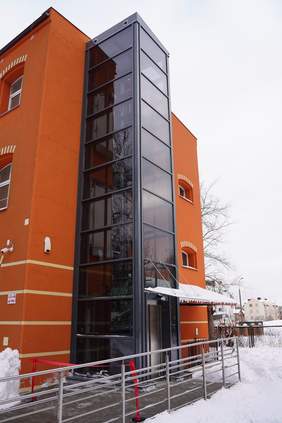 Pokaż zdjęcie: Ceglany budynek użyteczności publicznej z dobudowaną przeszkloną windą zewnętrzną. Przed wejściem metalowa rampa z poręczami, na ziemi śnieg. Winda łączy kilka kondygnacji i zapewnia dostęp osobom z ograniczoną mobilnością.