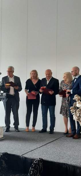 Pokaż zdjęcie: Sala konferencyjna w hotelu, na podwyższeniu stoi pięć osób, które trzymają w rękach dyplom i pamiątkowe serduszko.