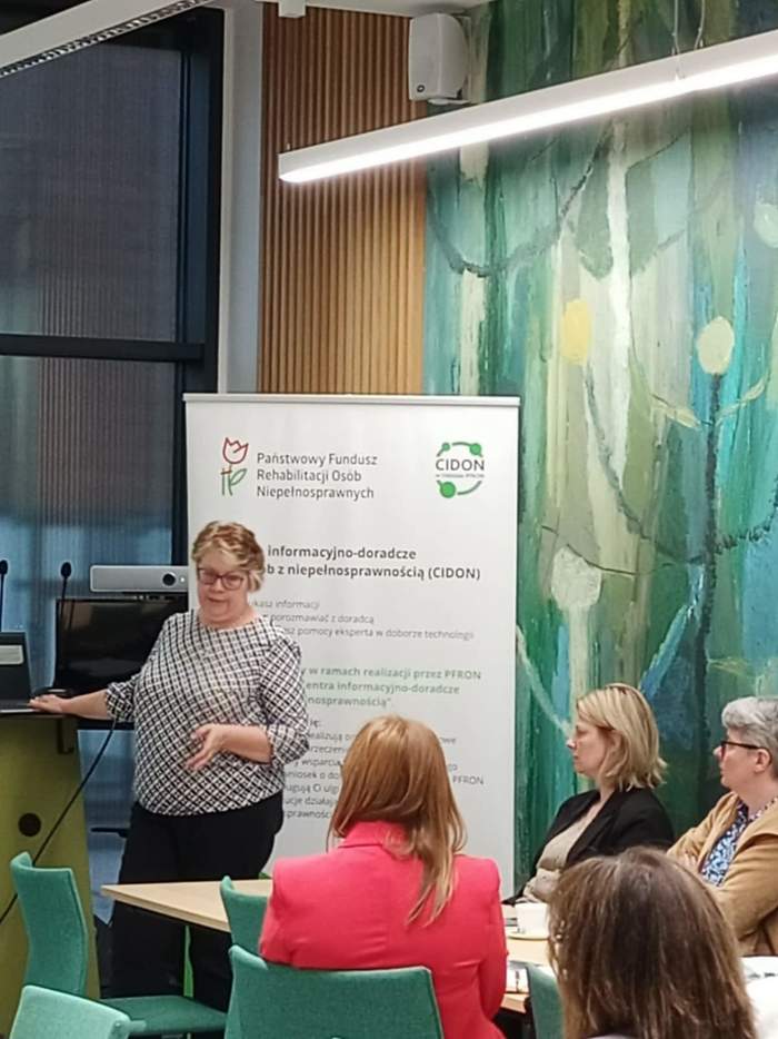 Kobiety siedzą przy stołąch i słuchają prelegentki z PFRON. Za nią stoi baner.