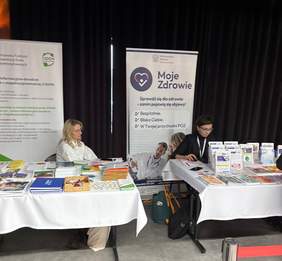 Pokaż zdjęcie: Stoisko informacyjne na wydarzeniu. Dwa stoły przykryte białymi obrusami, na nich rozłożone są różne materiały promocyjne oraz ulotki i broszury. Za stołami siedzą dwie kobiety – każda za osobnym stołem. Za nimi ustawione są dwa roll-upy z informacjami. Na lewym roll-upie widnieje logo Państwowego Funduszu Rehabilitacji Osób Niepełnosprawnych oraz napis Centrum informacyjno-doradcze dla osób z niepełnosprawnością. Na prawym roll-upie widnieje duże serce z napisem „Moje Zdrowie”.