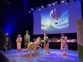 Pokaż zdjęcie: Aktorzy spektaklu w trakcie gry teatralnej, a za nimi wyświetlona prezentacja