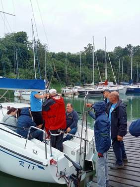 na pierwszym planie jacht, na nim kilka osób, na prawo poza jachtem na podeście również kilka osób, w tle widać jachty na jeziorze