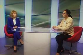 Pokaż zdjęcie: Przy szklanym stole siedzą dwie kobiety, zdjęcie robione z większej odległości, widać cale osoby. Kobieta po lewej stronie stołu, blondynka ubrana jest w niebieski garnitur, kobieta po prawej stronie szatynka, włosy spięte w kucyk, ubrana jest w beżową bluzkę i ciemne spodnie. 