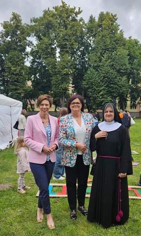 Pokaż zdjęcie: Trzy kobiety, w tym siostra zakonna stoją na trawniku. W tle bawiące się dzieci, fragment namiotu oraz drzewa liściaste.