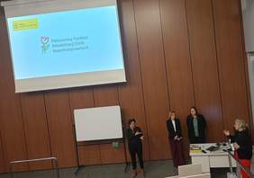 Pokaż zdjęcie: W sali wykładowej stoją cztery kobiety. Dwie z nich prowadzą szkolenie, trzecia zadaje pytania, a czwarta to tłumacz Polskiego Języka Migowego. W tle, na opuszczanym ekranie wyświetlane jest duże logo PFRON oraz małe logo „PFRON wspiera dostępność”.