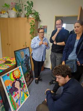 Pokaż zdjęcie: Cztery osoby w pokoju oglądają obrazy, jedna osoba kucnęła przed obrazem.