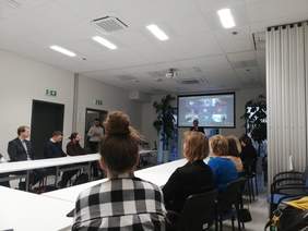 Pokaż zdjęcie: Widok na salę konferencyjną. Przy stołach ułożonych w literę U siedzą uczestnicy spotkania i patrzą na ekran i osobę prowadzącą spotkanie