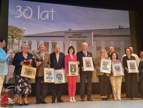 Pokaż zdjęcie: Na scenie kinoteatru stoją ludzie. Wszyscy trzymają w dłoniach obrazy z podziękowaniami za współpracę i wspieranie działań na rzecz środowiska osób z niepełnosprawnościami. 