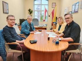 Przy podłużnym stole siedzi kobieta i trzech mężczyzn. Wszyscy patrzą w stronę aparatu. Zawierają umowę. Na stole leżą różne dokumenty. 