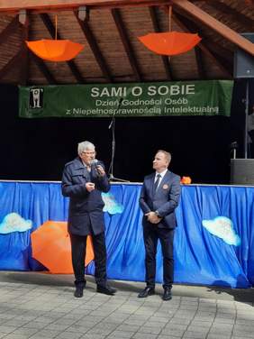 Pokaż zdjęcie: Przed sceną stoi dyrektor Łódzkiego Oddziału PFRON Władysław Skwarka. Trzyma w dłoniach mikrofon i przemawia. Obok stoi inny mężczyzna. 