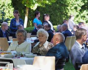 Pokaż zdjęcie: W plenerze przy stołach siedzą zaproszeni goście, kobiety i mężczyźni, stoły stoją w otoczeniu zieleni