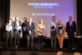 Pokaż zdjęcie: Grupowe zdjęcie wykonane na scenie, na której stoi osiem osób w eleganckich strojach. Osoby trzymają teczki z dokumentami, a za nimi na dużym ekranie widnieje napis: "RAZEM DLA BEZPIECZEŃSTWA - PREWENCJA 2024" oraz logo "Państwowa Inspekcja Pracy, Okręgowy Inspektorat Pracy w Szczecinie". To zdjęcie prawdopodobnie przedstawia moment wręczenia nagród lub wyróżnień związanych z inicjatywami dotyczącymi bezpieczeństwa w pracy.