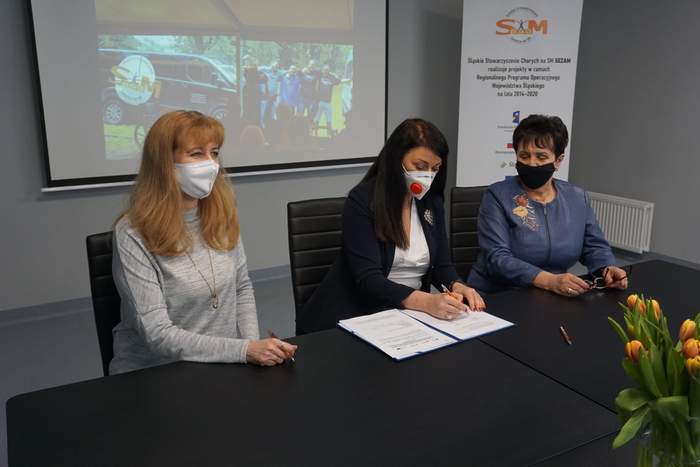 Pokaż zdjęcie: w pomieszczeniu siedzą trzy kobiety przy czarnym stole środkowa podpisuje dokumenty 