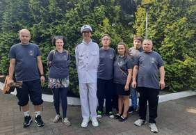 Pokaż zdjęcie: 6 osób pozuje do zdjęcia. Dwie kobiety i czterech mężczyzn. Po środku wysoki mężczyzna ubrany w biały strój żeglarza. Reszta osób w niebieskich koszulkach i ciemnych spodniach. W tle drzewa i ogród.  