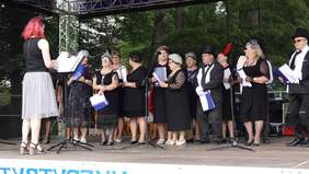 Na scenie grupa osób, seniorów, kobiet i mężczyzn w strojach w stylu lat 20tych, występują. Przed nimi kobieta, może dyrygent.