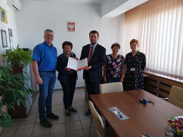 Pokaż zdjęcie: W gabinecie Dyrektora Oddziału PFRON w Zielonej Górze stoją obok siebie osoby podpisujące umowę, Dyrektor trzyma w ręku umowę, wszyscy są uśmiechnięci