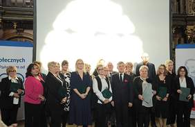 Pokaż zdjęcie: Grupa osób pozujących do wspólnej fotografii w trakcie oficjalnego wydarzenia. Na środku stoi mężczyzna w ciemnym garniturze z czerwoną poszetką, otoczony przez kobiety ubranych formalnie, w większości w odcieniach czerni, bieli oraz szarości. Niektóre z osób trzymają w rękach zielone teczki, co sugeruje, że mogły otrzymać wyróżnienia lub dokumenty. W tle znajduje się duży ekran projekcyjny, na którym światło tworzy jasną, rozmytą plamę. Po bokach widoczne są banery związane z Zakładem Ubezpieczeń Społecznych, co wskazuje na oficjalny charakter spotkania. Wnętrze, bogato zdobione, nadaje wydarzeniu uroczysty nastrój.