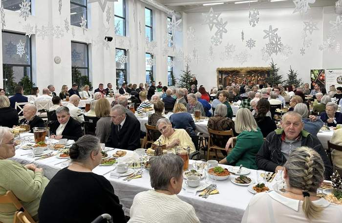 Pokaż zdjęcie: Zdjęcie sali w której przy zastawionych stołach siedzą pensjonariusze domu pomocy społecznej oraz goście. Z sufitu zwisają gwiazdki i śnieżynki.