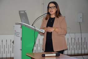 Pokaż zdjęcie: Przy mównicy z logo PFRON stoi kobieta w beżowej marynarce.